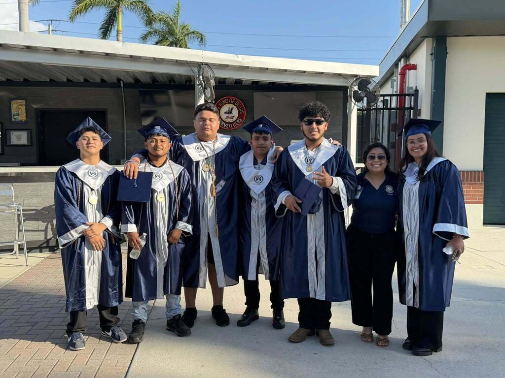 Six high school graduates smile wearing navy gowns while standing with a staff member.
