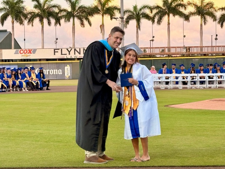 A school principal in a black gown, congratulate a graduate, wearing a white gown by handing off a diploma
