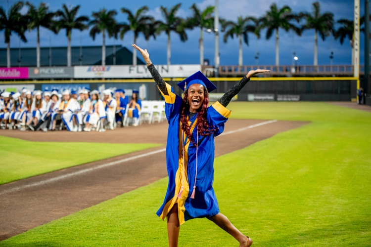 A high school graduate in a blue and gold gown, holds her hand up in the air to celebrate