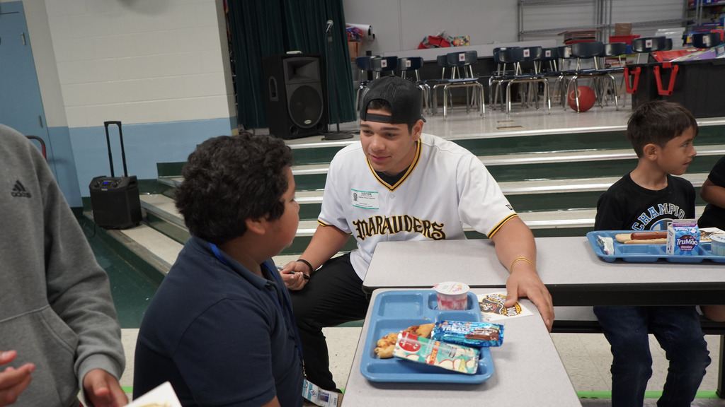 A minor league baseball player sits with a boy whose eating with a breakfast tray in front of him.