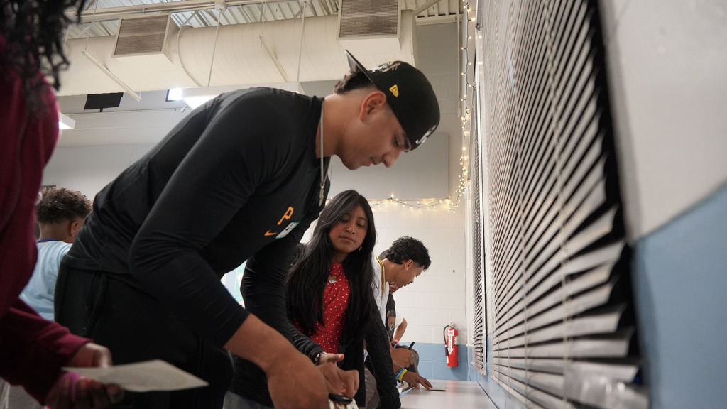 A baseball player leaning over while signing an autograph as a student watches on.