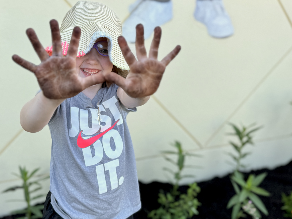 A little girl wearing a Nike shirt that reads "Just Do It" holds up her soil covered hands after working in the garden.