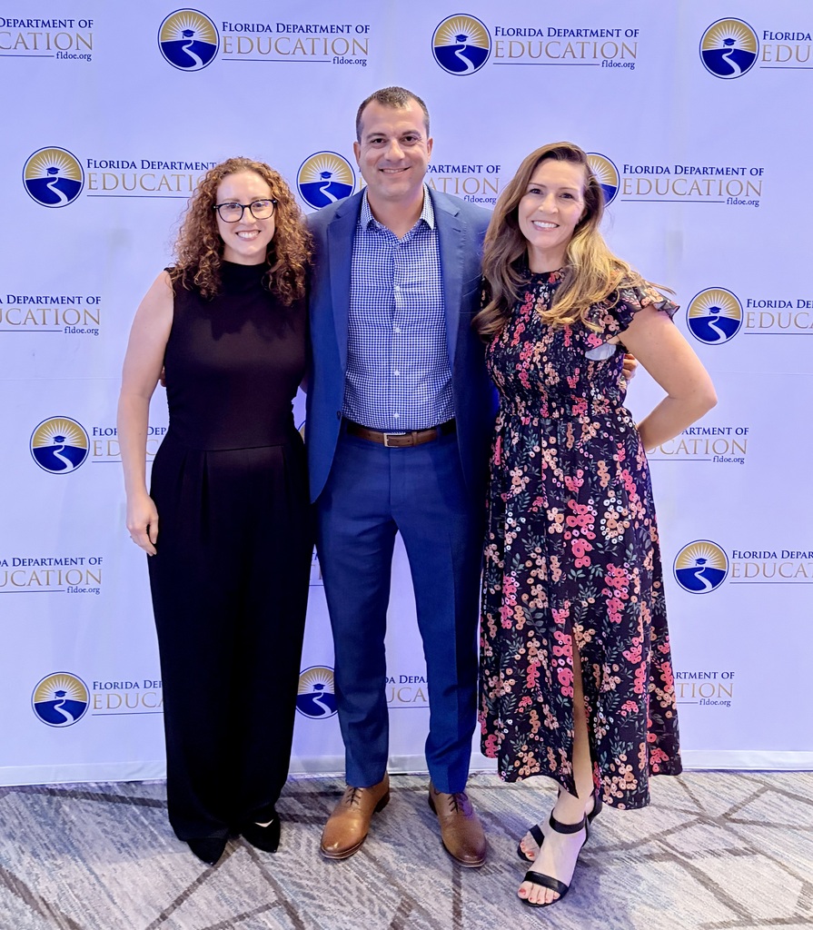 Three smiling adults standing in front of a backdrop with logos for the Florida Department of Education