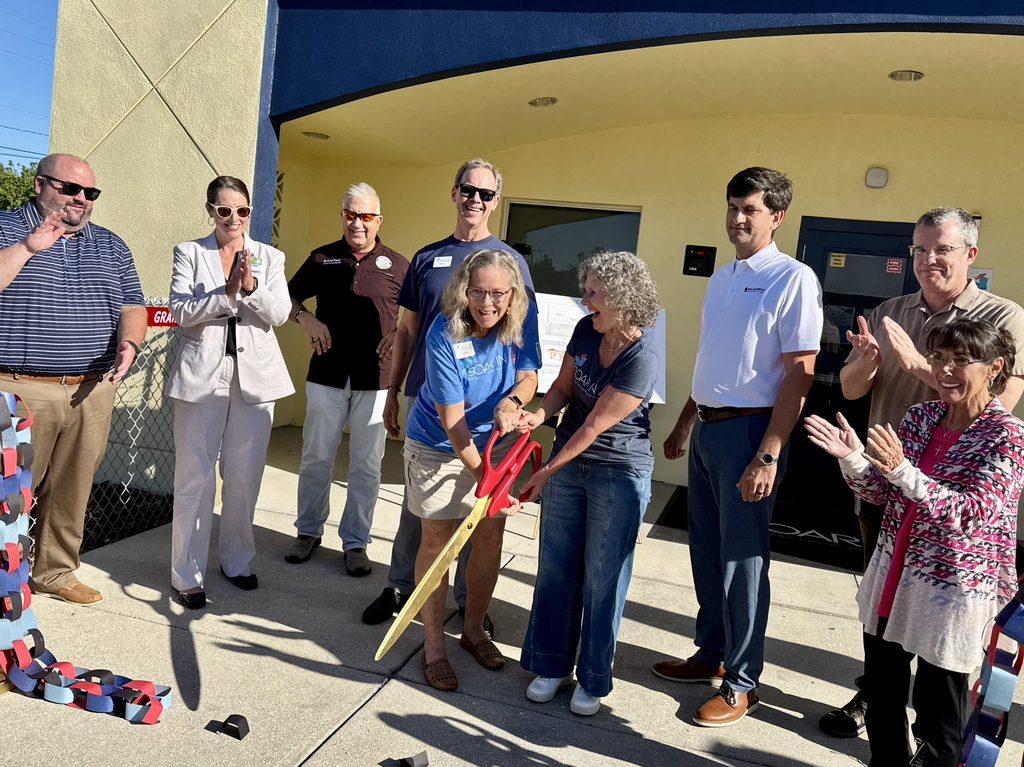 A group of people smile and clap as two women cut a paper chain 'ribbon' with a giant pair of gold scissors.