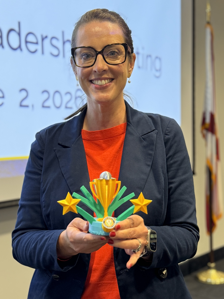 A woman in red shirt and blue jacket holds a 3D printed trophy featuring gold, green and blue. 
