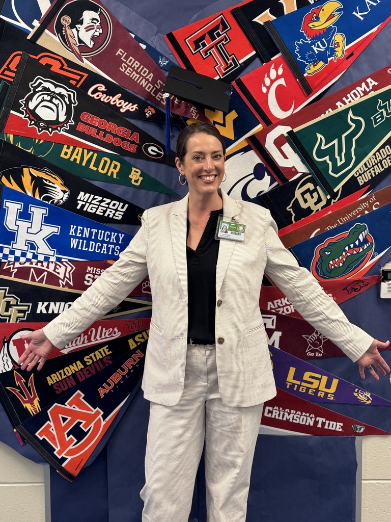 A woman wearing a suit holds out her arms while standing in front of a mural in which college pennants make a pair of wings
