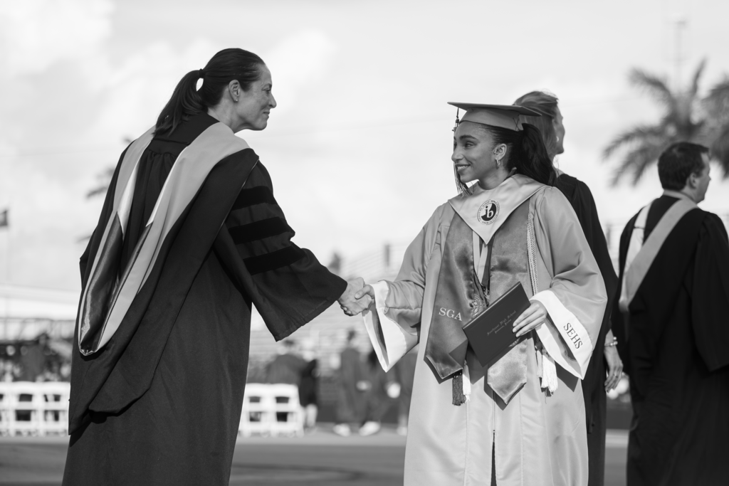 A black and white photo of a woman and teen both dressed in graduation gowns shaking hands.
