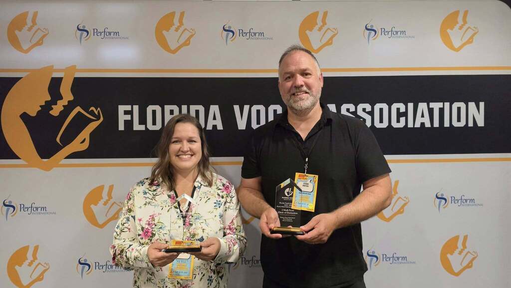 A female and male teacher holding glass trophies in front of a backdrop for Florida Vocal Association.