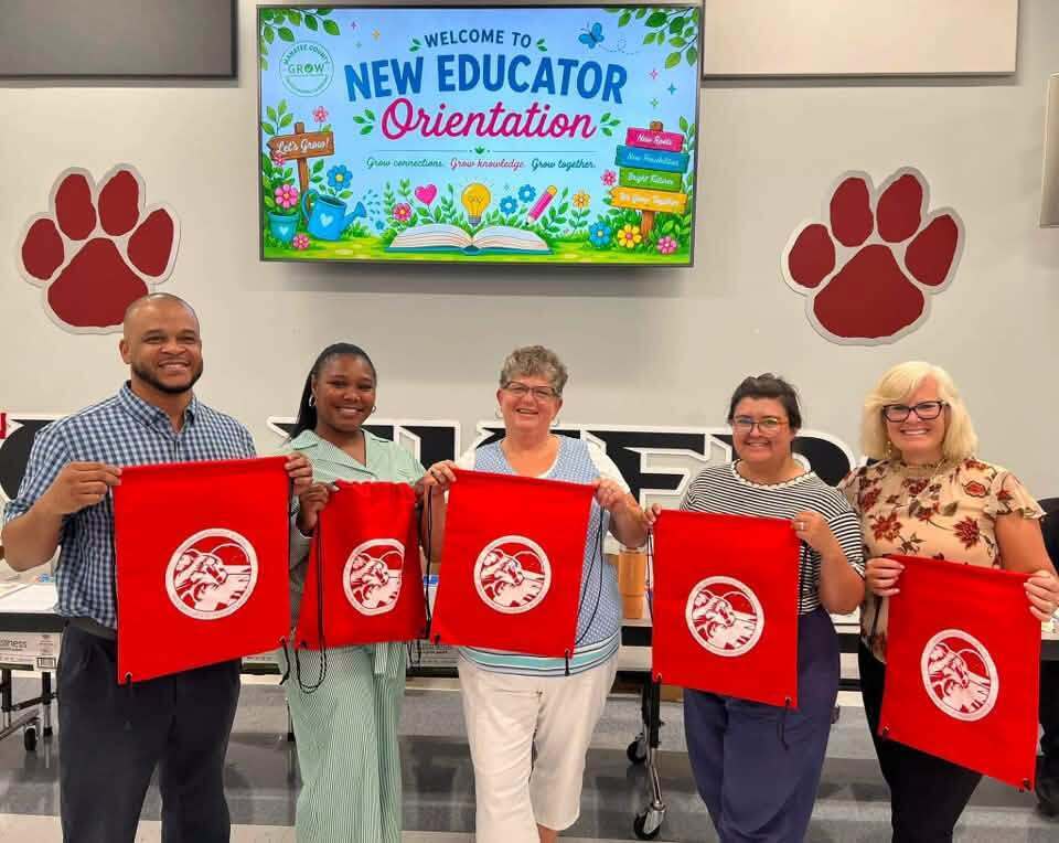 Smiling teachers holding red drawstring bags under a television showing a graphic that says "Welcome to New Educator Orientation"