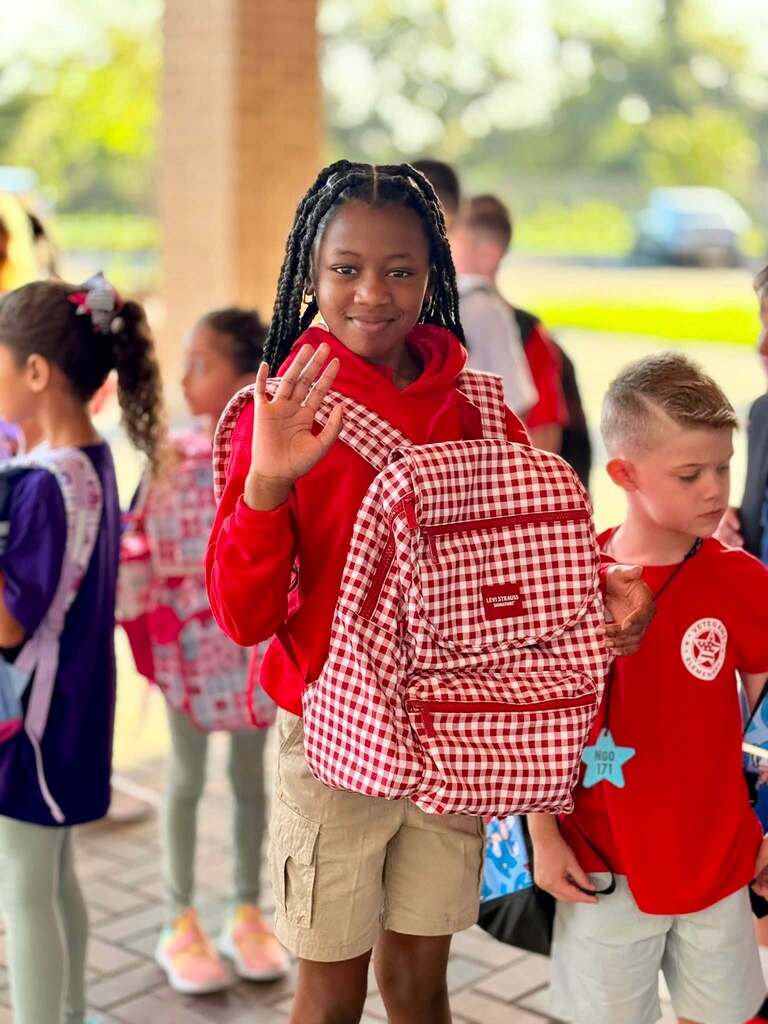A young woman waves while wearing a red gingham backpack.