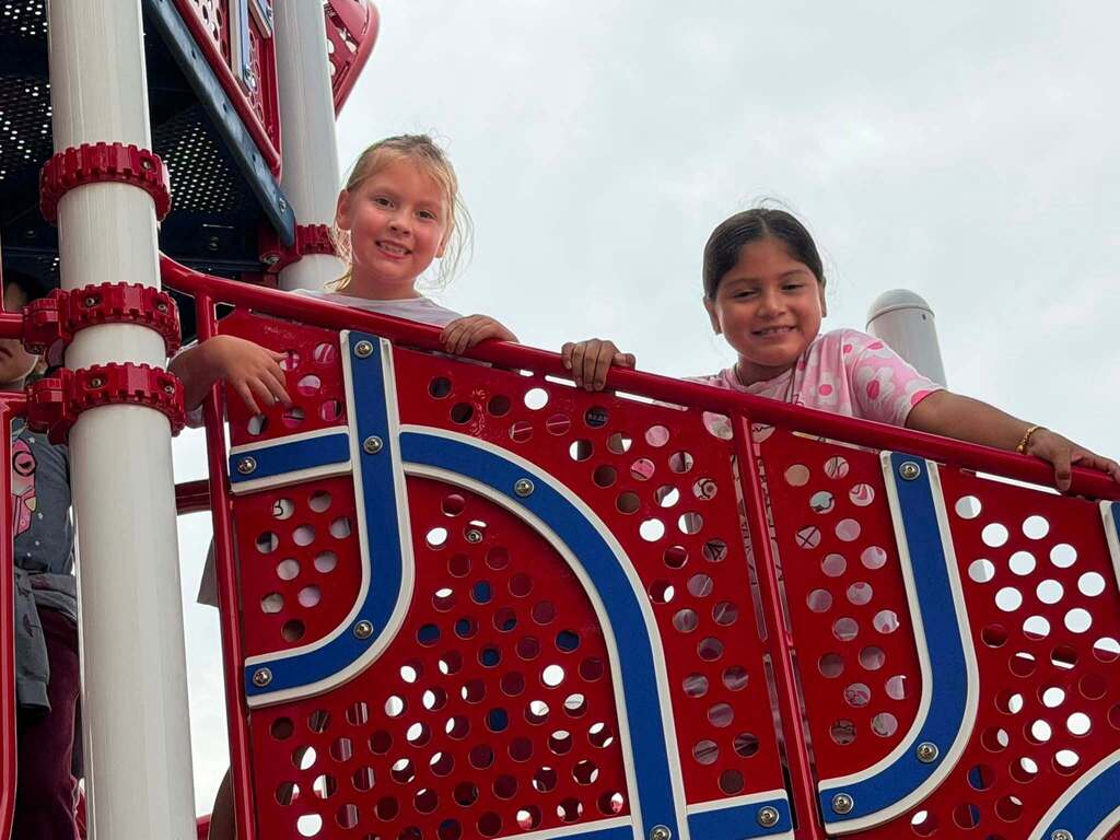 Two girls smiling while standing on red, white, and blue playground equipment.