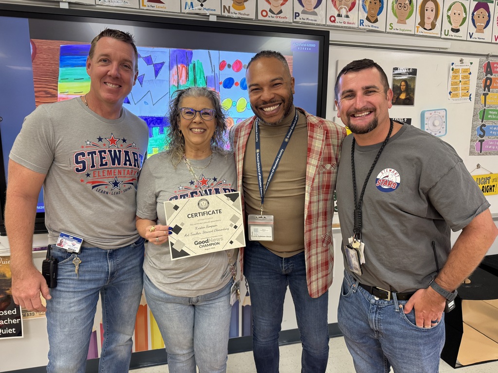 Four smiling adults stand shoulder to shoulder in an elementary school art classroom.