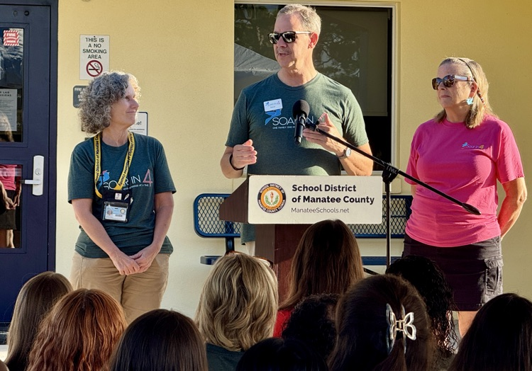 A man speaks at a podium as two women stand next to him