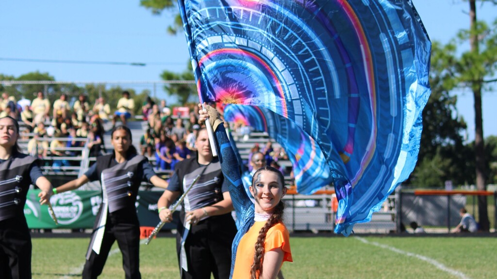 A colorguard performer wearing an orange and blue uniform smiles while hoisting a flag in an outdoor stadium setting.