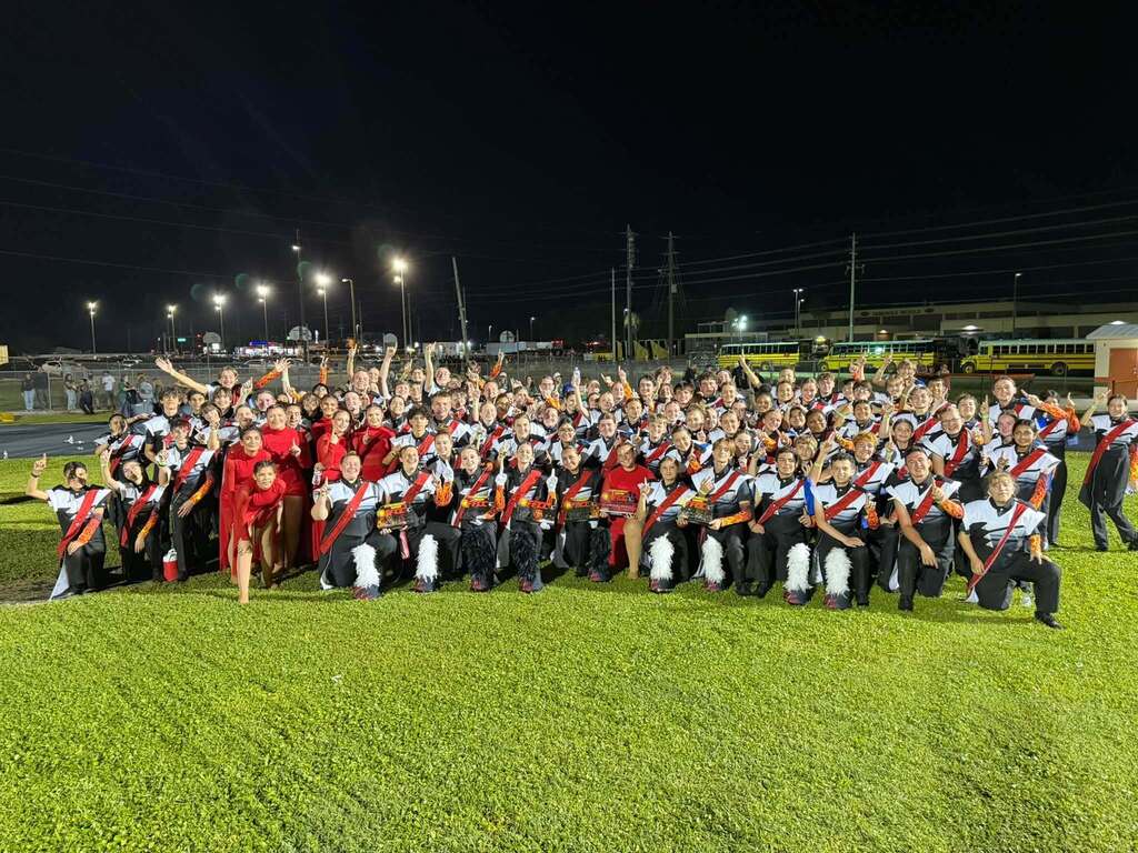 Dozens of high school marching band performers smiling while wearing colorful uniforms on green grass.