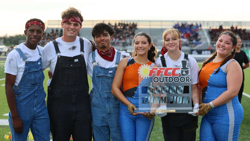 Teens wearing overalls and colorful uniforms holding a Grand Champion trophy.