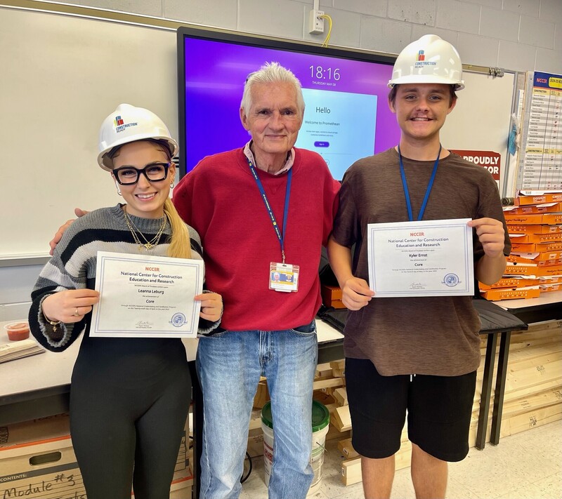 Two students in construction hats holding certifications with their teacher smiling