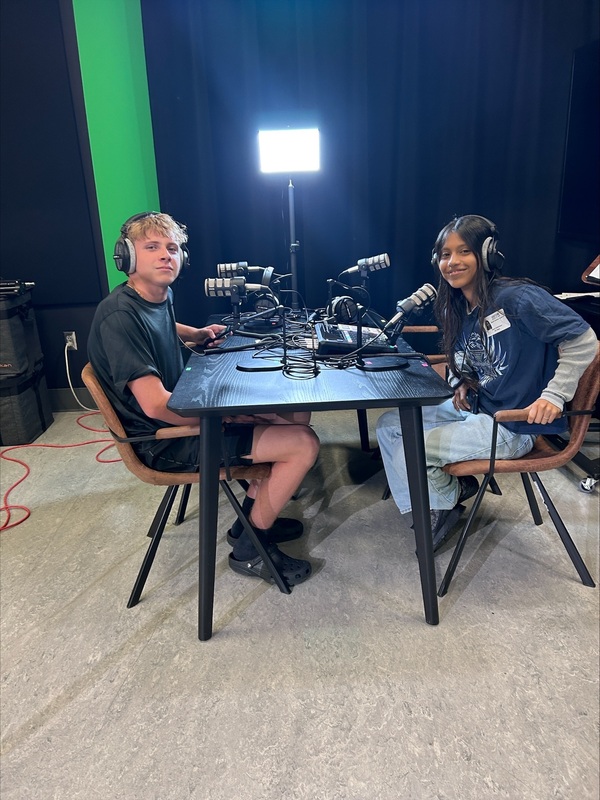 Two students wearing headphones sit across from each other at a table with microphones.