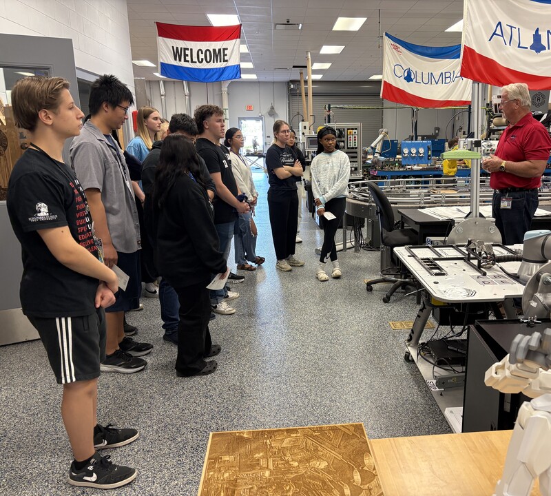 Students visiting a manufacturing class with flags hanging from the ceiling that say Welcome.