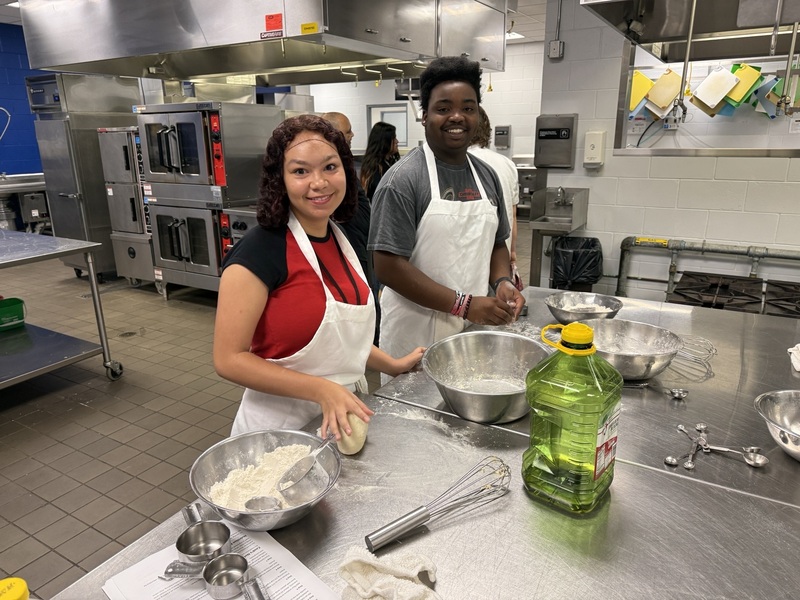 A young man and young woman wearing hairnets and white aprons smile in a kitchen and cooking lab.