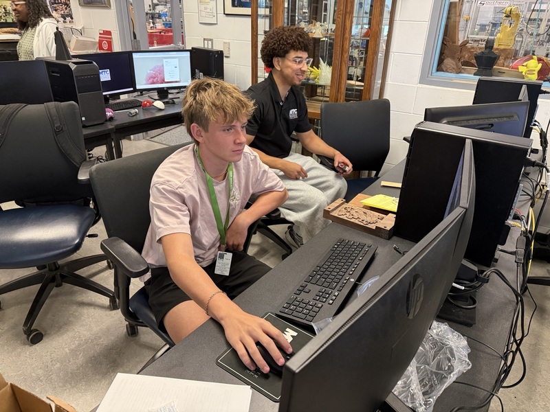 A college student and high school student sit on computers in a lab setting.