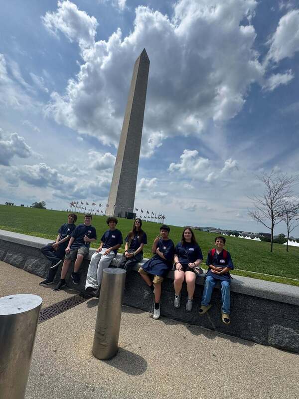 A group of teens sitting on a wall in front of a tall monument with blue sky and clouds behind them.