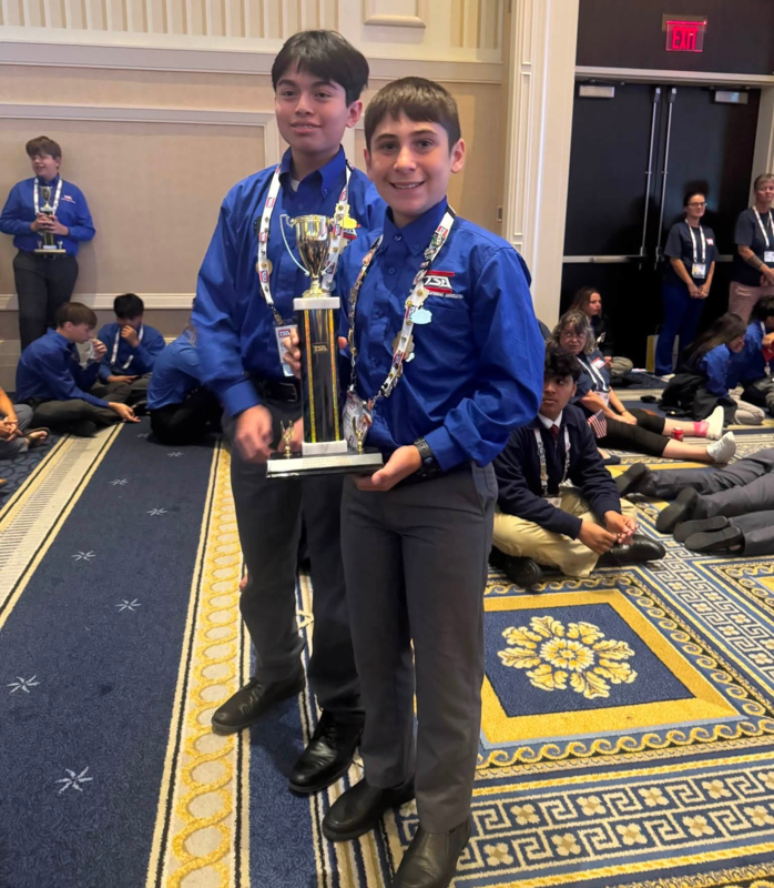 Two students in long sleeve blue dress shirts holding a trophy and smiling.
