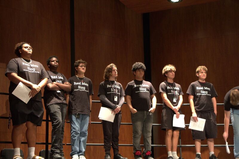 Young men wearing black shirts which read, "Real Men Sing" stand shoulder to shoulder on stage.