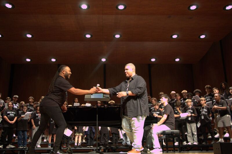 A man wearing all black hands a microphone to another man wearing a black shirt and khaki slacks.