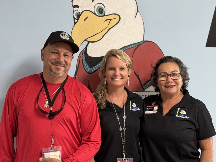 three smiling members of a school staff standing in front of a wall with an eagle paint on it