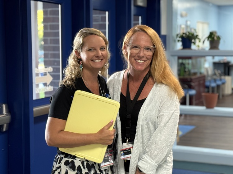 A school principal holding a clipboard smiles alongside the school’s media specialist