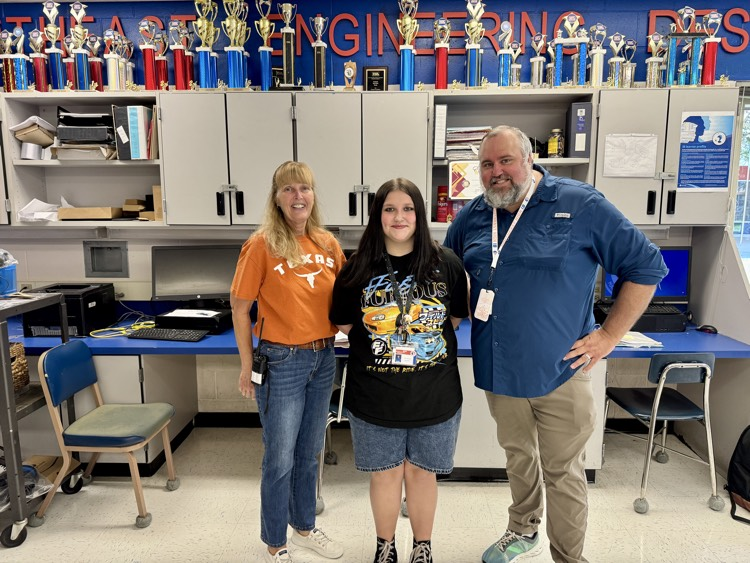 two adult school staff members surround a student with trophies, lining the wall of a classroom behind them