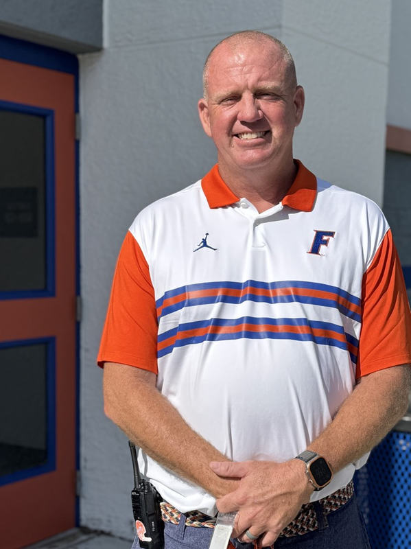 A man standing in front of a school building, wearing an orange blue and white polo shirt