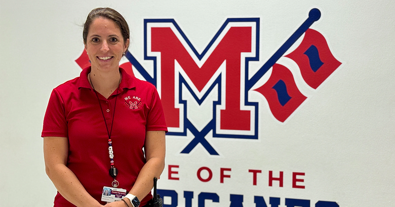 A smiling principal stands in front of the school logo painted on a wall