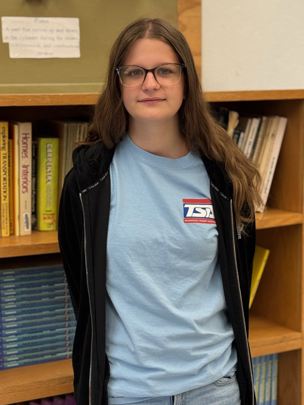 A teenager wearing a TSA tshirt and black jacket standing in front of a book shelf.