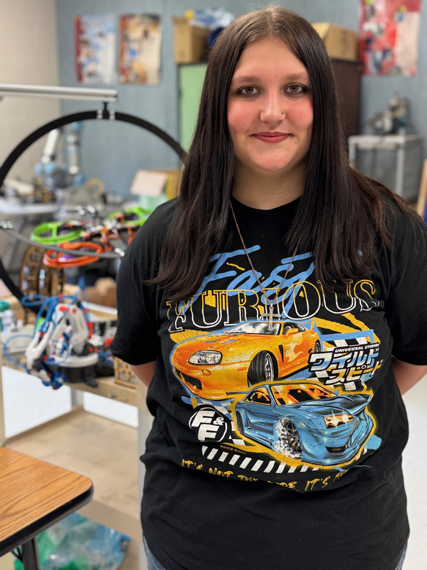 A teenager wearing a black tshirt with cars on it stands in an engineering lab at a high school.