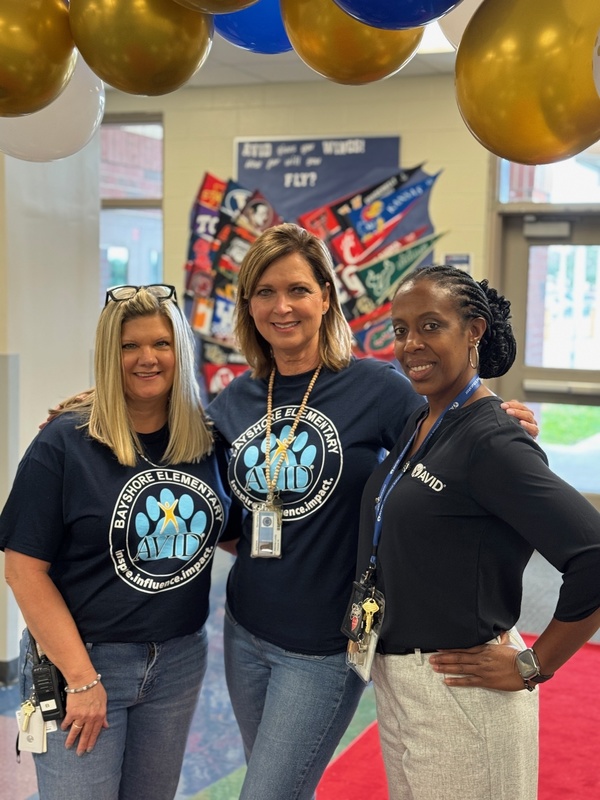 Three women smiling under a balloon arch inside a school.