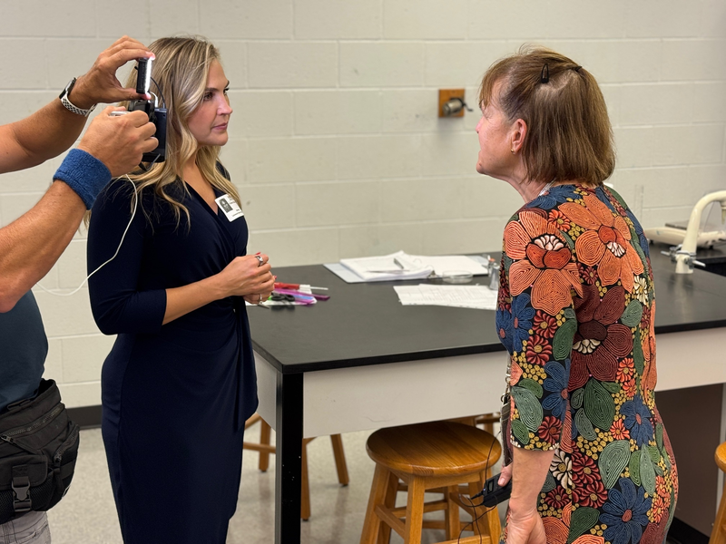 A TV news reporter interviews a teacher wearing a floral dress.