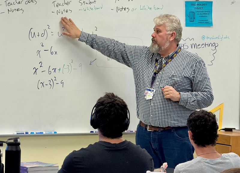 A math teacher points to an equation on a classroom white board.