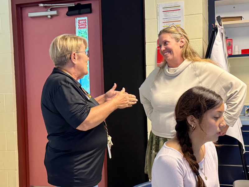 Two colleagues talking in a classroom with a student nearby doing work on a computer.