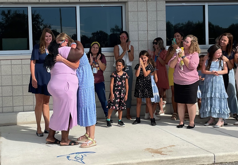 Woman in pink dress being hugged by classroom teacher she works with every day.