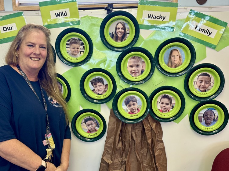 A pair of professionals stands next to a bulletin board with photos of all her students