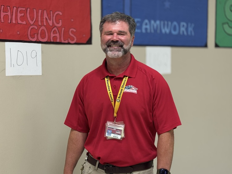 A school principal wearing a red polo shirt smiles in the hallways