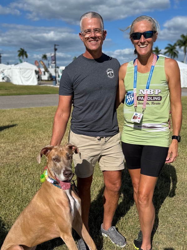 A man, woman, and great dane dog with blue skies and clouds in the sky.