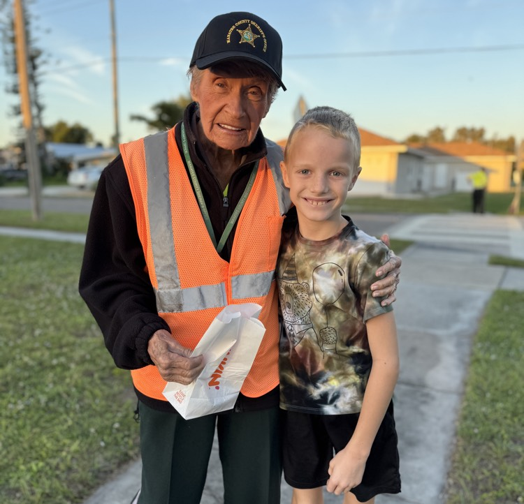 A boy and a tie-dye T-shirt delivers his school crossing guard a doughnut for her birthday