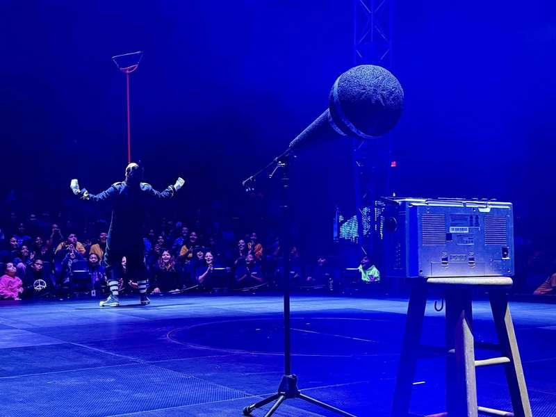 Performer Johnny Rockett balances a broom on his chin as an audience of children laugh.