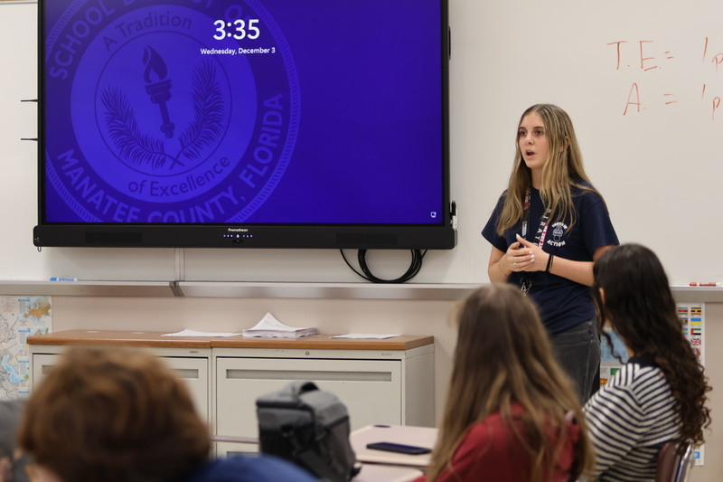 a girl speaks in front of a class of teens.