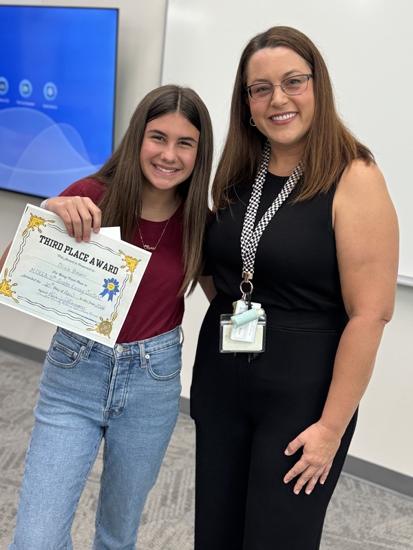 A young girl in a red tshirt and jeans stands beside her teacher and smiling.