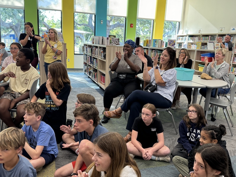 A media center filled with teachers, students, and parents, celebrate a winning essay contest.
