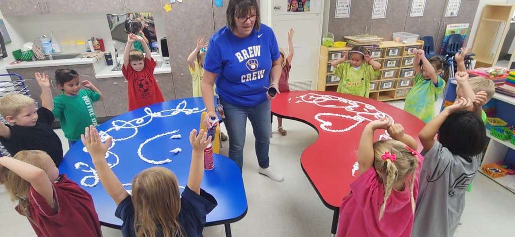 Early elementary students standing around bean shaped tables with their hands in the air. The teacher is between the tables, spreading shaving cream on them, which the students will explore.